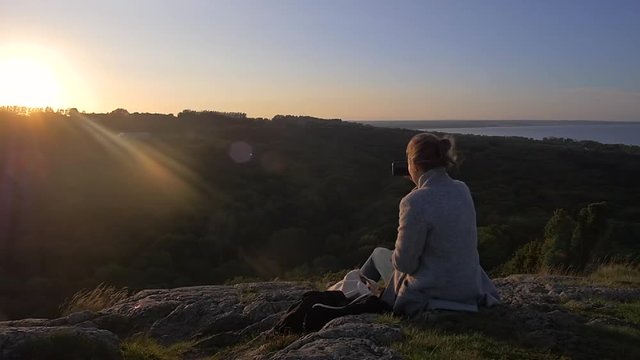 Woman watching Sunset after Hike on Top of Mountain while taking Pictures Overlooking the Baltic Sea.