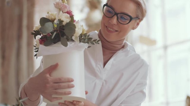 Tilt up shot of cheerful middle aged female florist examining beautiful flower composition in hatbox, then looking at camera and happily smiling