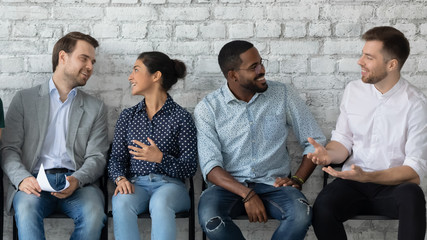 Happy motivated young multiethnic people sit in queue line talk waiting for office interview, smiling diverse multiracial job candidates have fun chatting before recruitment talk, employment concept