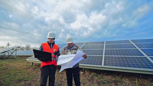 Solar panels installation. Two workers on the background of blue photovoltaic panel. Engineers in hard hat looking into the laptop and scheme plan on a solar farm.