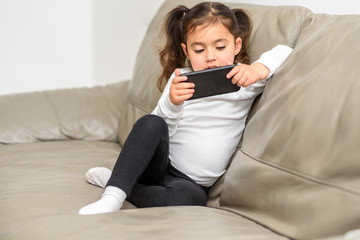 Beautiful Small Girl Attending To Online Class From Home. Little Child With Cell Phone On Couch.