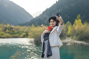 Fototapeta premium Beautiful young mother with her infant baby in sling make selfie. Mother is carrying her child and travel in autumn mountain lake. Active mother concept