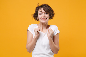 Excited young brunette woman girl in white t-shirt posing isolated on yellow orange background studio portrait. People lifestyle concept. Mock up copy space. Clenching fists, wait for special moment.