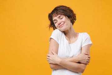 Obraz premium Smiling young brunette woman girl in white t-shirt posing isolated on yellow orange background. People lifestyle concept. Mock up copy space. Keeping eyes closed holding hands crossed hugging herself.