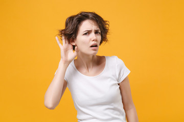 Curious young brunette woman in white t-shirt posing isolated on yellow orange background studio...