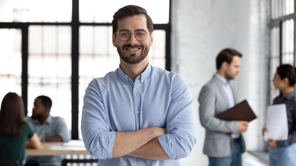 Smiling young Caucasian businessman in glasses stand in modern office show confidence and motivation at work, happy millennial European male CEO posing at workplace, leadership, success concept - obrazy, fototapety, plakaty