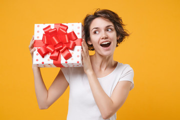 Curious young woman in white t-shirt posing isolated on yellow orange background. Valentine's Day Women's Day birthday holiday concept. Mock up copy space. Hold red present box with gift ribbon bow.