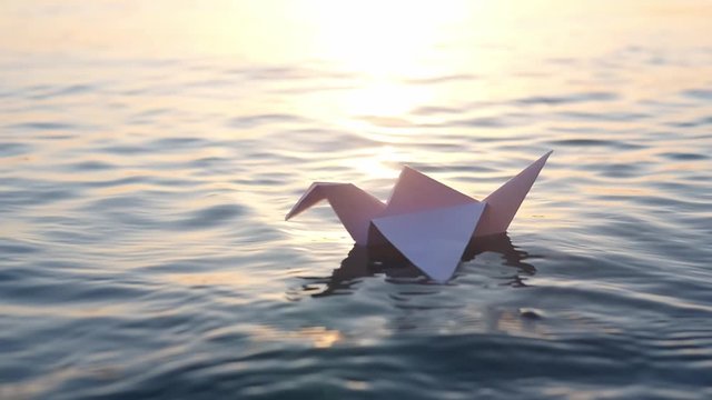 A paper crane floats through the waves in the water at a beautiful sunset. Origami crane saving lives. The concept of a dream, future, childhood, freedom or hope. Slow motion. Sasaki Origami Sadako