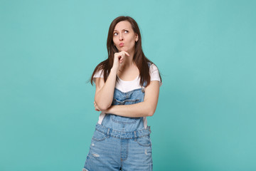 Pensive confused young brunette woman girl in casual denim clothes posing isolated on blue turquoise background. People lifestyle concept. Mock up copy space. Put hand prop up on chin, looking up.