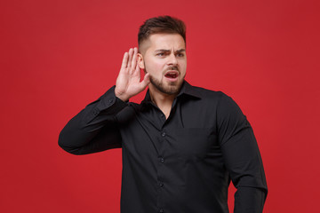 Shocked young bearded guy in classic black shirt posing isolated on red background studio portrait....