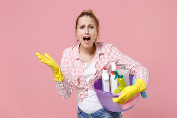 Shocked worried young woman housewife in rubber gloves hold basin with detergent bottles washing cleansers doing housework isolated on pink background in studio. Housekeeping concept. Spreading hands.