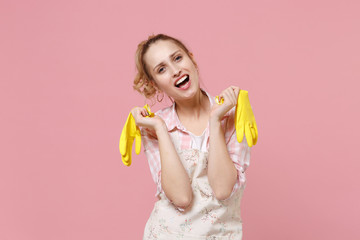 Cheerful laughing young woman housewife in casual clothes, apron doing housework isolated on pastel pink wall background studio portrait. Housekeeping concept. Hold in hands yellow rubber gloves.