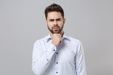 Puzzled young unshaven business man in light shirt posing isolated on grey wall background studio portrait. Achievement career wealth business concept. Mock up copy space. Put hand prop up on chin.