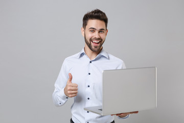 Cheerful young unshaven business man in light shirt isolated on grey wall background. Achievement career wealth business concept. Mock up copy space. Working on laptop pc computer, showing thumb up.