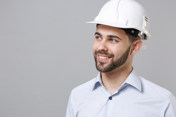 Smiling young unshaven business man in light shirt protective construction helmet posing isolated on grey background. Achievement career wealth business concept. Mock up copy space. Looking aside.