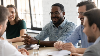Happy multiracial young businesspeople sit at desk in office laugh brainstorm discuss business...