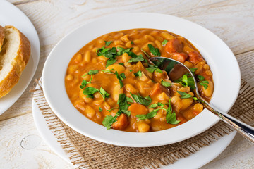 White beans in tomato sauce with sausage slices and parsley in a white bowl and spoon on a wood table. Bean is source of vegetable protein and ingredient for vegetarian food.