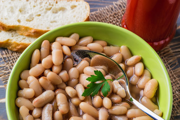 White backed beans and a spoon in a green bowl near slices of bread on a table. Bean is source of vegetable protein and ingredient for vegetarian food.