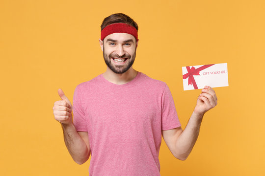 Smiling young bearded fitness sporty guy sportsman in headband t-shirt in home gym isolated on yellow background. Workout sport motivation lifestyle concept. Hold gift certificate, showing thumb up.
