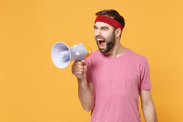 Crazy young bearded fitness sporty guy 20s sportsman in headband t-shirt spend weekend in home gym isolated on yellow background. Workout sport motivation concept. Scream in megaphone, looking aside.
