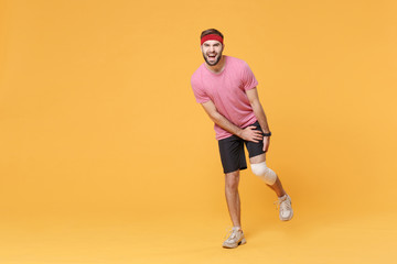 Fototapeta na wymiar Ingured displeased young bearded fitness guy 20s sportsman in headband t-shirt in home gym isolated on yellow wall background. Workout sport motivation concept. Touch leg with elastic bandage on knee.