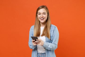 Fototapeta premium Cheerful young woman girl in casual denim clothes posing isolated on bright orange wall background studio portrait. People lifestyle concept. Mock up copy space. Using mobile phone typing sms message.