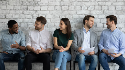 Fototapeta premium Happy young multiracial interns or applicants have fun talking before office interview, smiling multiethnic job candidates sit in queue line chatting before recruitment talk, employment concept