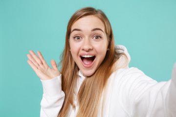 Close up of excited young woman girl in casual white hoodie isolated on blue turquoise background. People lifestyle concept. Mock up copy space. Doing selfie shot on mobile phone, spreading hands.