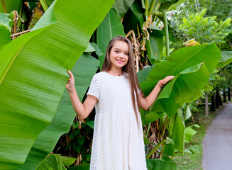 Portrait of young, smile model girl in white dress in banana palm leaves. Cute summer portrait. Fashion, pretty, teenage girl with long hair in outdoors. Warm summer day and vacation time.