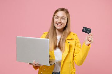 Naklejka premium Smiling young woman girl in yellow leather jacket posing isolated on pastel pink background studio portrait. People lifestyle concept. Mock up copy space. Hold laptop pc computer, credit bank card.