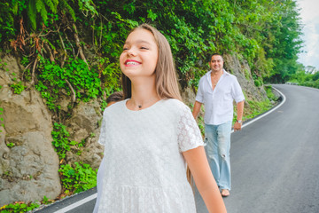 Cheerful, happy daughter enjoying walk play outdoor. Funny family in white clothes barefoot running on walkway. Green rocks at background.