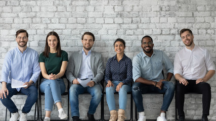 Portrait of smiling diverse multiracial young people sit in row on chairs wait for office...