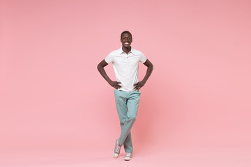 Fototapeta na wymiar Smiling young african american man guy in white polo shirt, turquoise trousers posing isolated on pastel pink wall background. People lifestyle concept. Mock up copy space. Standing with arms akimbo.