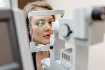 Young beautiful woman with blond hair is diagnosed with eye pressure on special ophthalmological equipment - obrazy, fototapety, plakaty