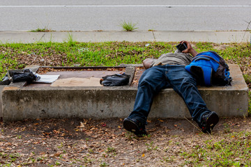 Homeless man with a modern cell phone taking a break  laying down on a piece of concrete next to the road