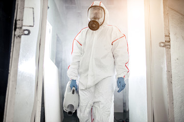 professional disinfector in protective suit holding chemical sprayer and other equipment for...