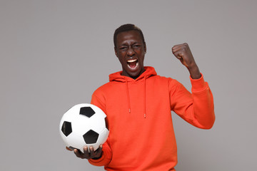 Screaming young african american man guy football player in orange streetwear hoodie isolated on grey background. Sport leisure lifestyle concept. Play football hold soccer ball doing winner gesture.