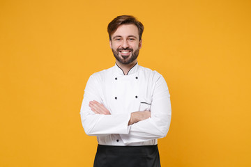 Smiling young bearded male chef cook or baker man in white uniform shirt posing isolated on yellow orange background studio portrait. Cooking food concept. Mock up copy space. Holding hands crossed. - obrazy, fototapety, plakaty