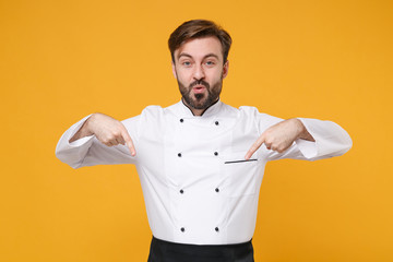 Amazed young bearded male chef cook or baker man in white uniform shirt posing isolated on yellow background studio portrait. Cooking food concept. Mock up copy space. Pointing index fingers down.