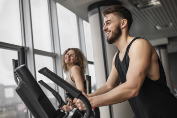 Fototapeta na wymiar Handsome fitness instructor is running with his attractive client how to work out on an exercise bike in gym