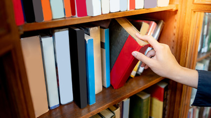 Male university student hand choosing and picking old book from old wooden bookshelf in college...
