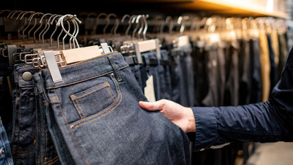 Male hand customer choosing black men jeans or denim pants (trousers) hanging on rack in clothes shop. Fashion product collection in clothing store for selling. Textile industry and business concept - obrazy, fototapety, plakaty