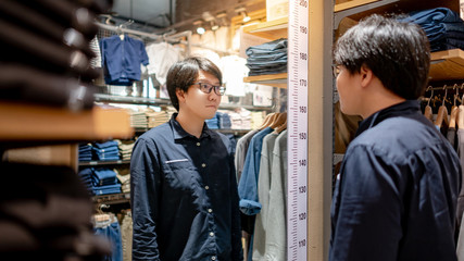 Asian man customer looking at himself in the mirror in while shopping new cotton shirt in clothing store. Choosing and trying casual clothes in fitting room of cloth shop. Urban lifestyle concept