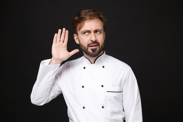 Curious young bearded male chef cook or baker man in white uniform shirt posing isolated on black background in studio. Cooking food concept. Mock up copy space. Try to hear you with hand near ear.
