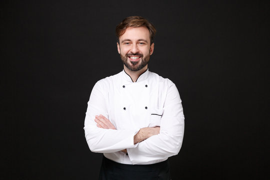 Smiling young bearded male chef cook or baker man in white uniform shirt posing isolated on black wall background studio portrait. Cooking food concept. Mock up copy space. Holding hands crossed.