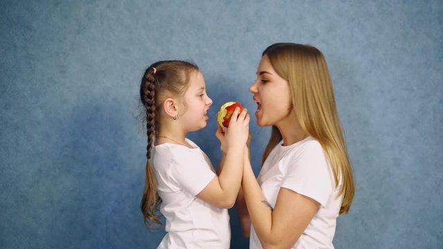 Little girl and her mother in white t-shirts bite an apple. Side view of mother and daughter eating one ripe apple on blue studio background.