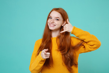 Smiling young redhead girl in yellow sweater isolated on blue turquoise background. People lifestyle concept. Mock up copy space. Doing phone gesture says call me back pointing index finger on camera.