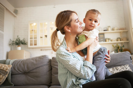 Indoor image of cute young female with ponytail holding tight her charming baby, sitting on sofa with him. Pretty mother and son bonding in living room, mom looking at child with love and tenderness