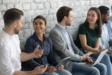 Smiling diverse multiethnic young people sit in line have fun talking waiting for interview, happy...