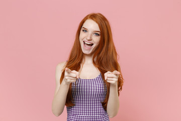 Cheerful young redhead woman girl in plaid dress posing isolated on pastel pink background studio portrait. People emotions lifestyle concept. Mock up copy space. Pointing index fingers on camera.